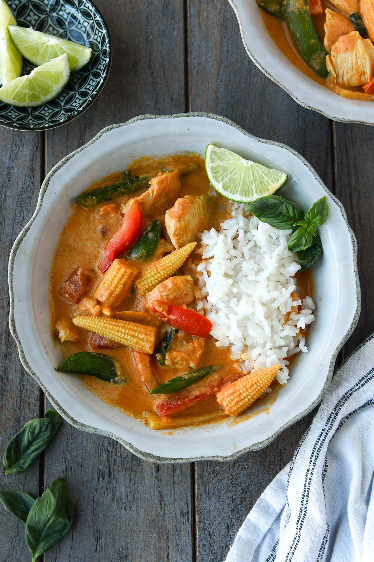 Above view of a bowl of Thai red curry served with rice, a sprig of of basil and a wedge of lime. A striped towel, basil leaves, and a small bowl of lime slices are around the bowl.