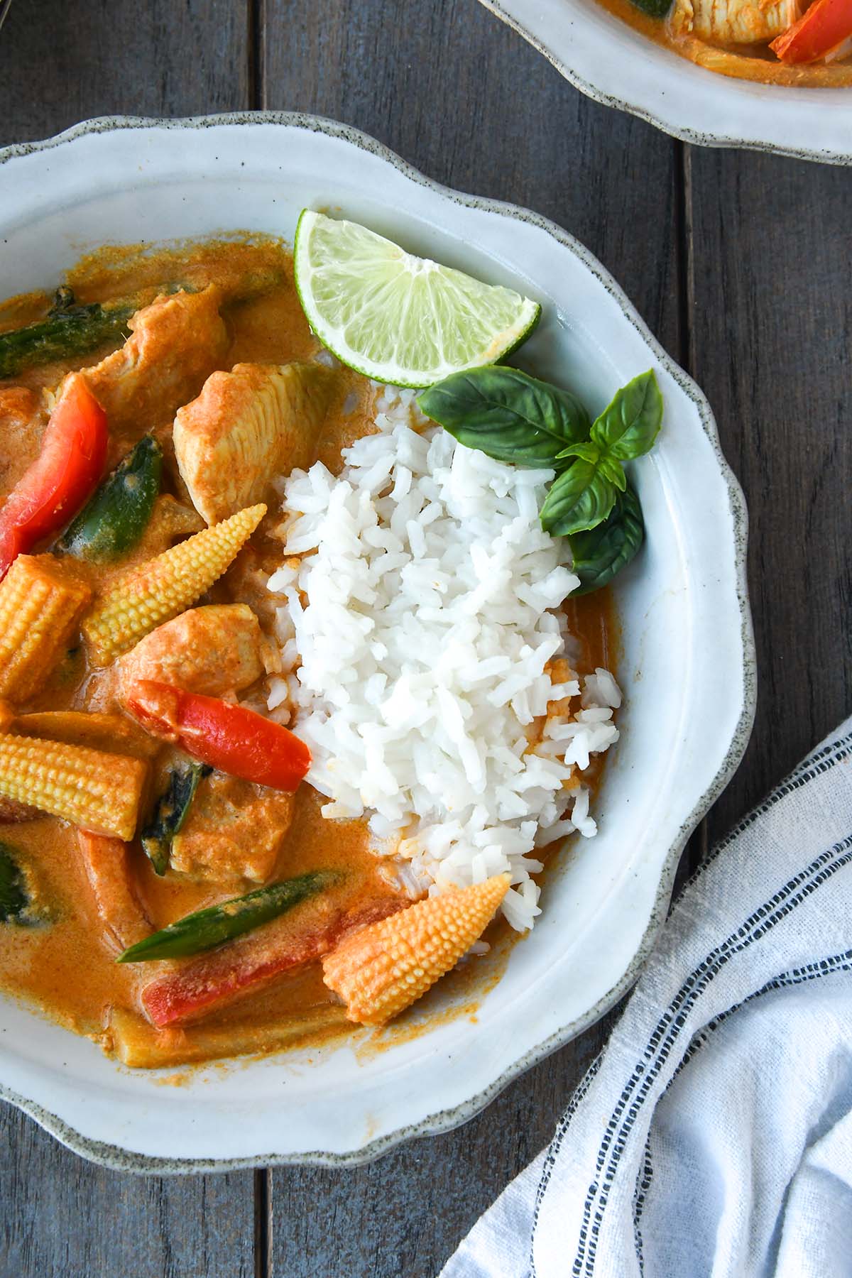 Up close above view of a bowl of Thai red curry served with rice, a sprig of of basil and a wedge of lime. A striped towel and another bowl of curry next to the bowl.