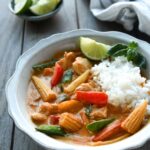 A bowl of Thai red curry served with rice, a sprig of of basil and a wedge of lime.