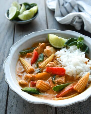 A bowl of Thai red curry served with rice, a sprig of of basil and a wedge of lime.