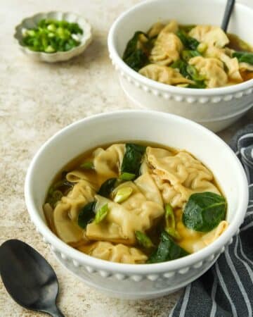 Two bowls of wonton soup with a small bowl of sliced green onions. A spoon and a towel.