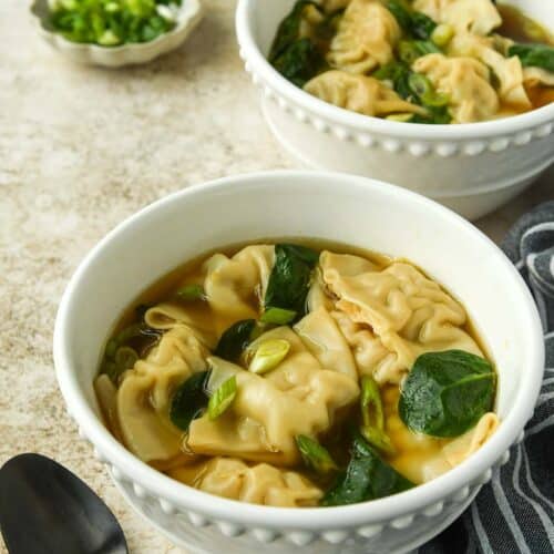 Two bowls of wonton soup with a small bowl of sliced green onions. A spoon and a towel.