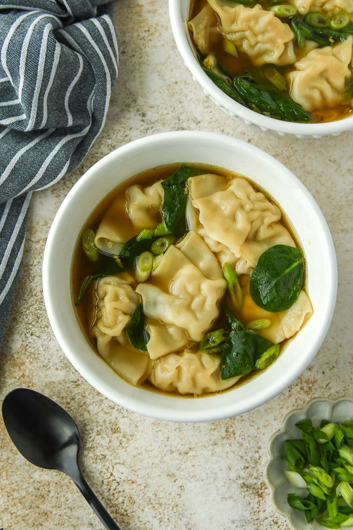 Above view of a bowl of wonton soup with cut onions and spinach leaves. A spoon and a striped towel are nearby.