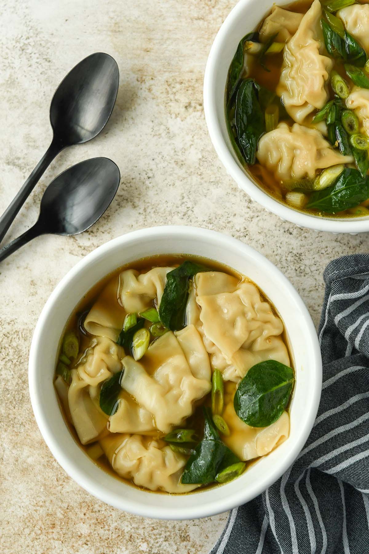 Above view of a bowl of wonton soup with cut onions and spinach leaves. Two spoons and a striped towel.