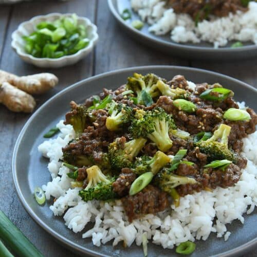 Two plates of ground beef and broccoli topped with sliced green onions and sesame seeds. With a small dish of green onions, a piece of fresh ginger and a striped towel.