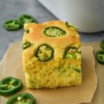 A square of cornbread on parchment with a pan of cornbread and some sliced jalapeños.