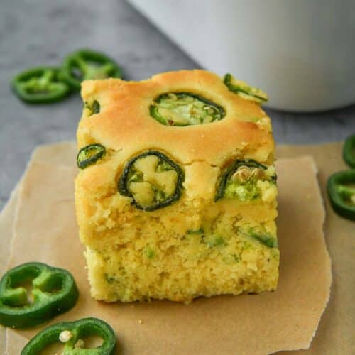 A square of cornbread on parchment with a pan of cornbread and some sliced jalapeños.