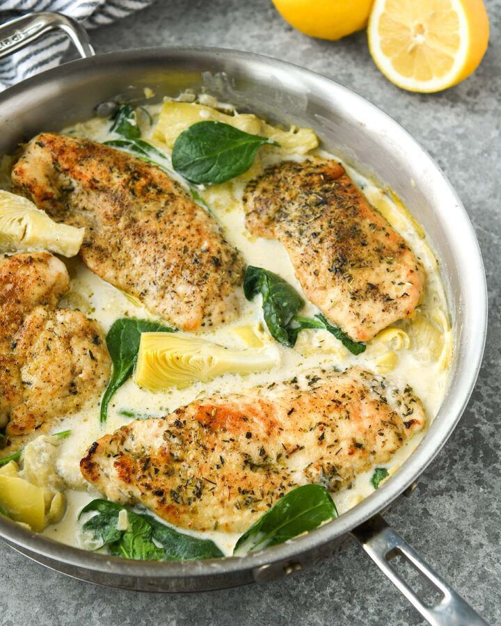 Above view of spinach artichoke chicken in a pan with a striped towel and lemon halves.