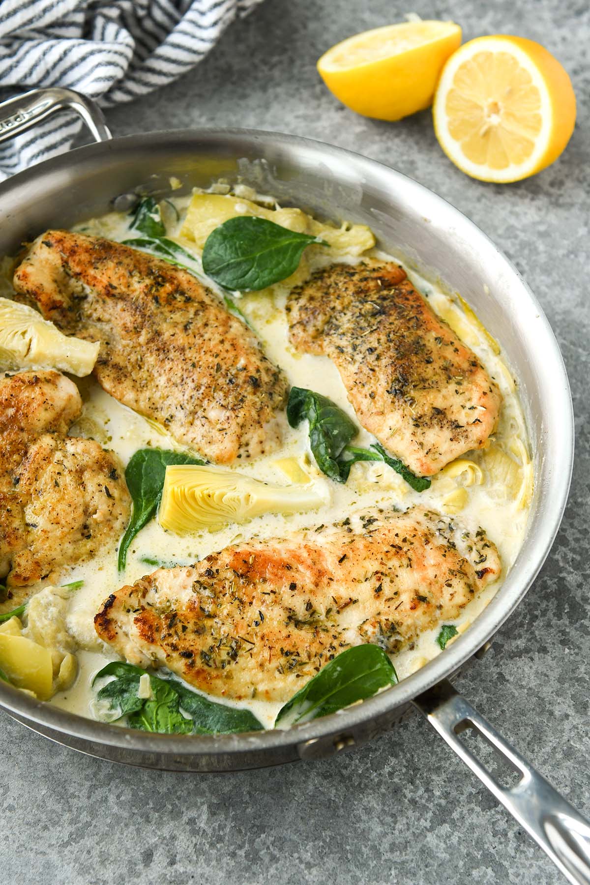 Above view of spinach artichoke chicken in a pan with a striped towel and lemon halves.