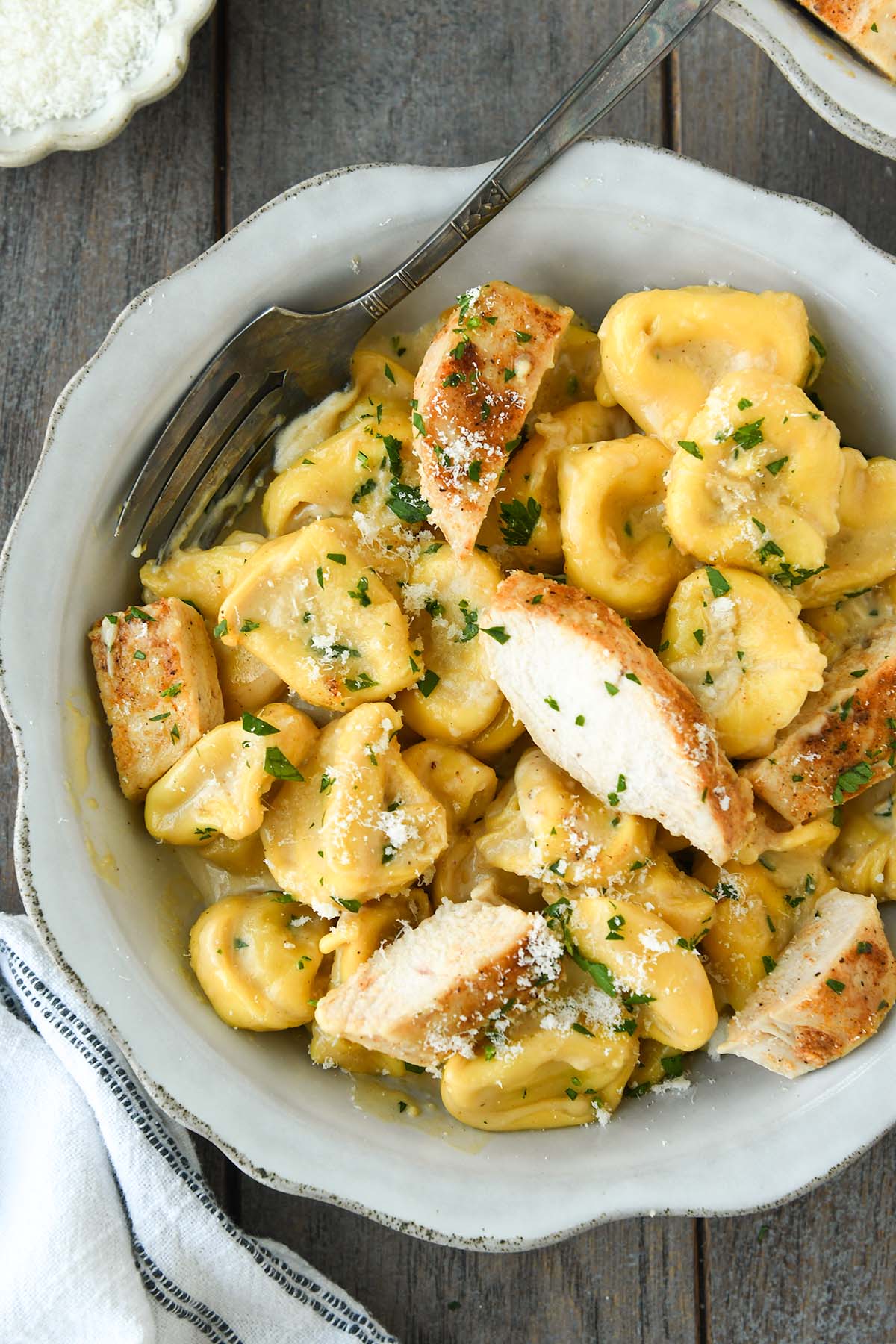 Above view  and up close of a bowl of tortellini alfredo with chicken chunks, a small bowl of parmesan and a fork.