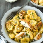Above view of a bowl of tortellini alfredo with chicken chunks, a small bowl of parmesan and a white striped towel.