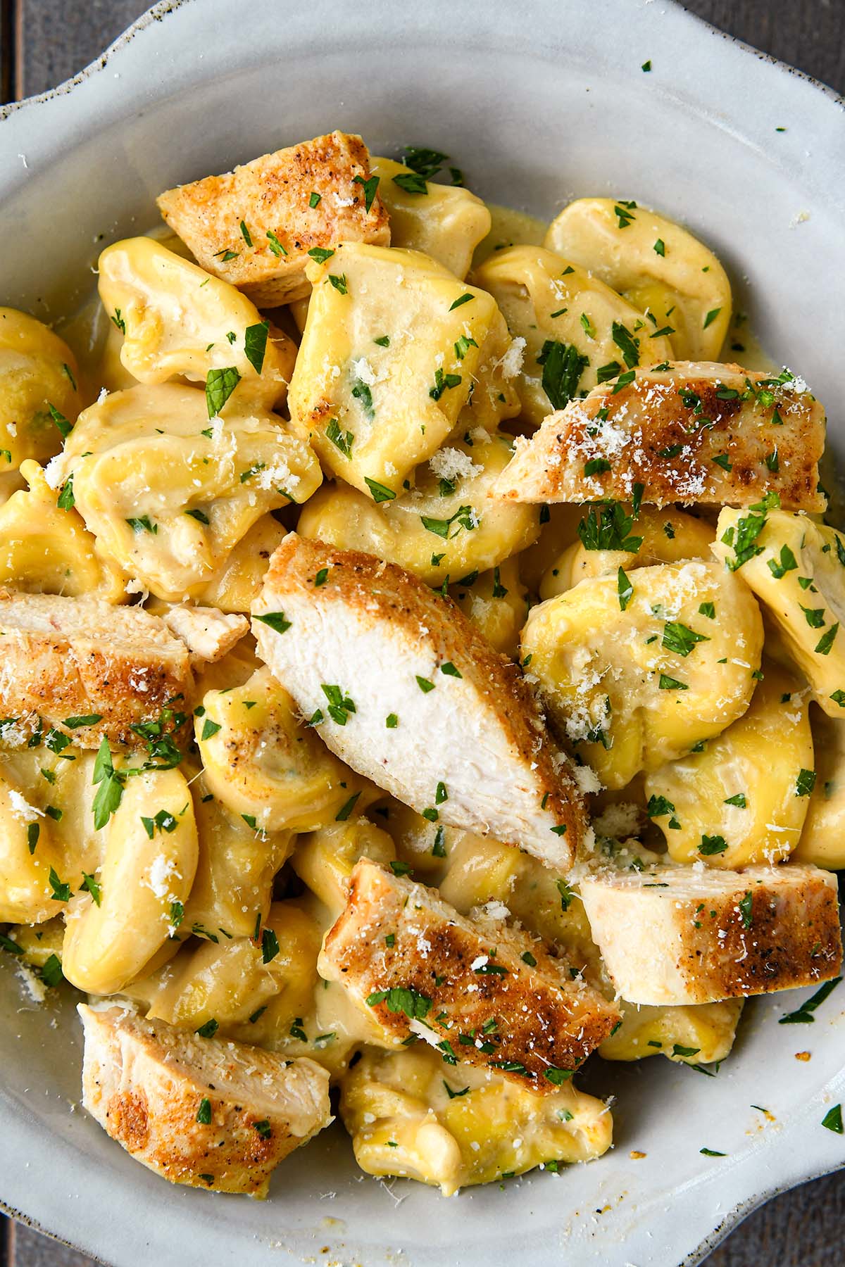 Super up close of a bowl of tortellini alfredo with chicken chunks, a small bowl of parmesan and a fork.