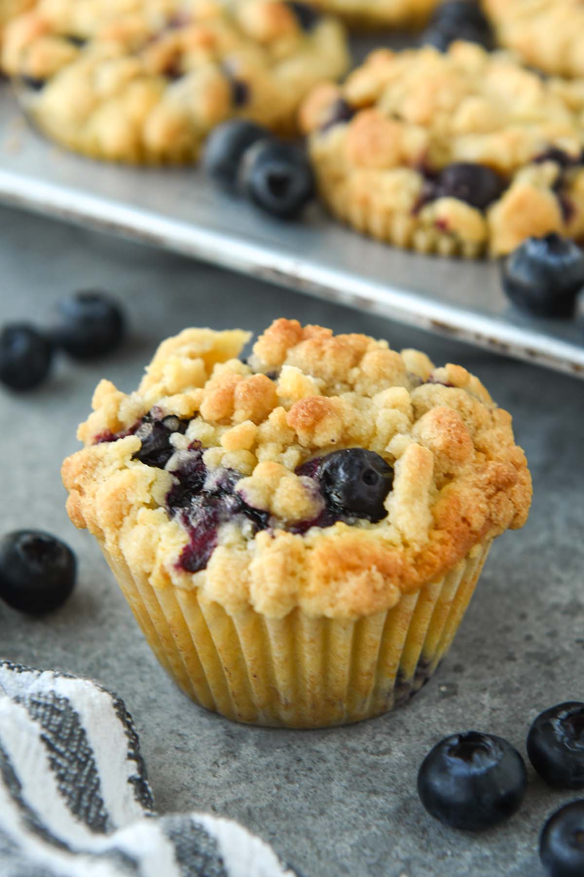 Up close of a blueberry muffin with some fresh blueberries around and a striped towel. There a tin of muffins in the background.