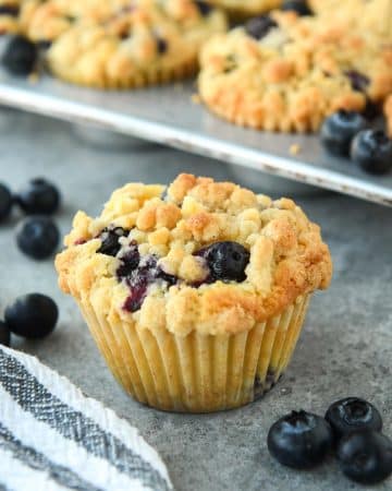 Up close of a blueberry muffin with some fresh blueberries around and a striped towel. There a tin of muffins in the background.