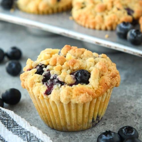 Up close of a blueberry muffin with some fresh blueberries around and a striped towel. There a tin of muffins in the background.