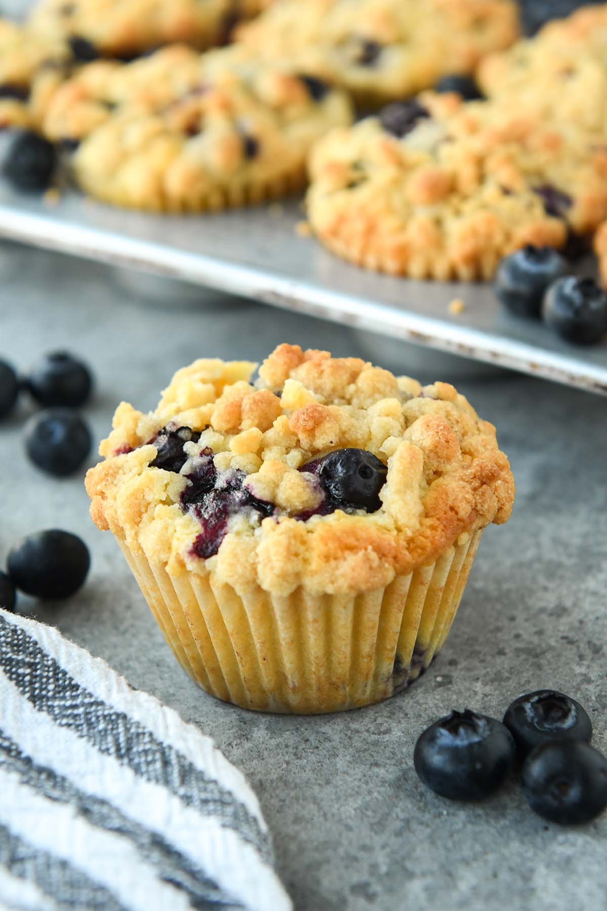 Up close of a blueberry muffin with some fresh blueberries around and a striped towel. There a tin of muffins in the background.