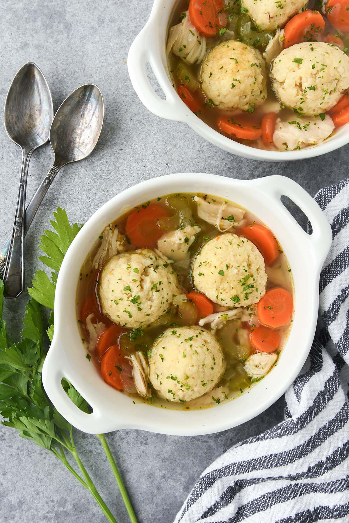 Above view of matzo ball soup with chicken and vegetables, a striped towell and spoons.
