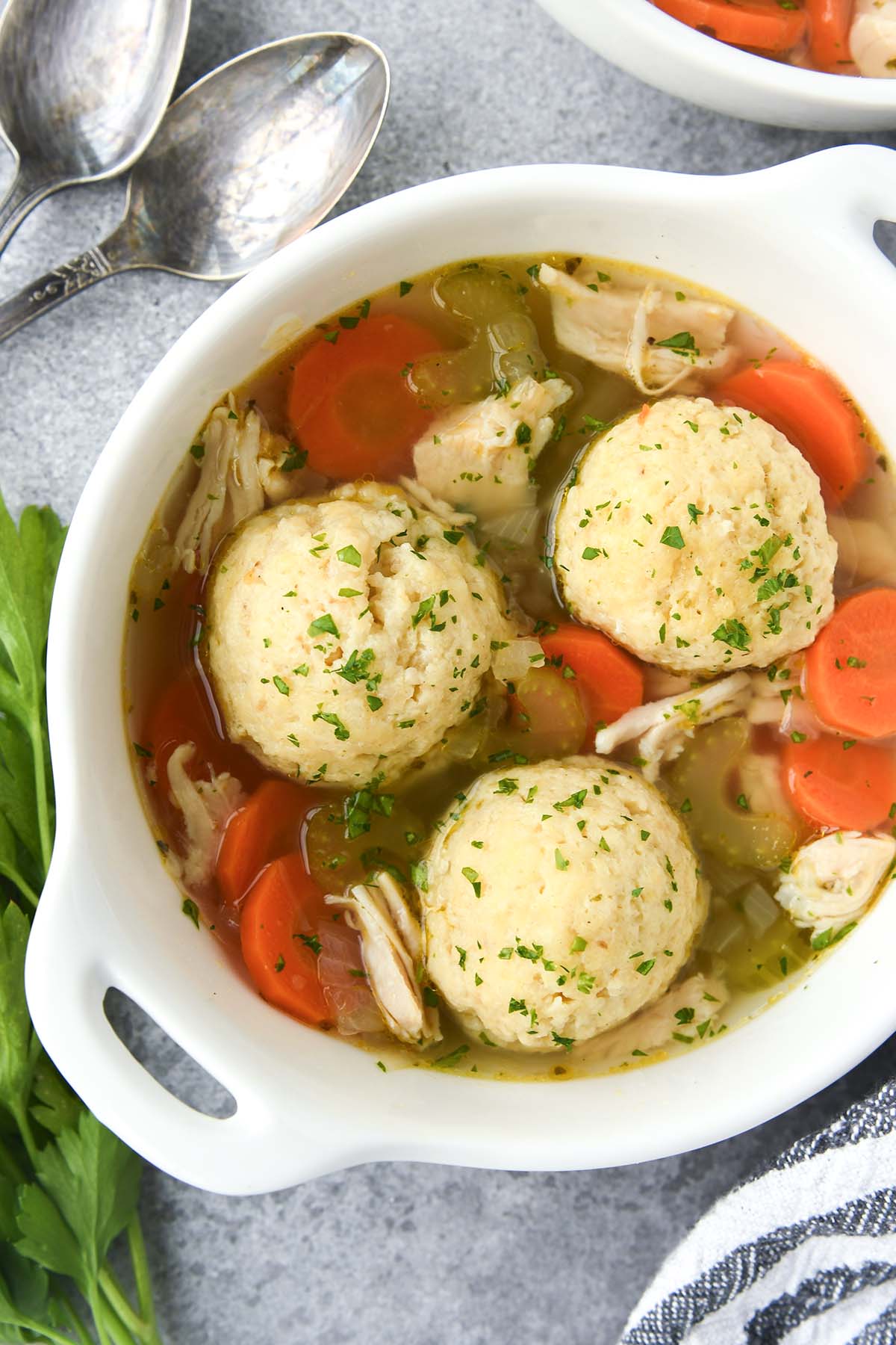 Above view of matzo ball soup with chicken and vegetables, a striped towell and spoons.