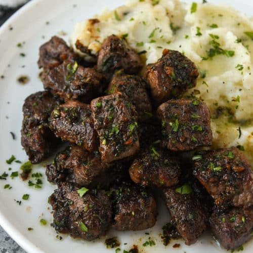 A plate of garlic steak bites sprinkeld with chopped parsley with mashed potatoes.