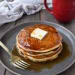 A plate with a stack of packcakes with a pat of butter and drizzeld with syrup. A red coffee cup and a towel in the background.