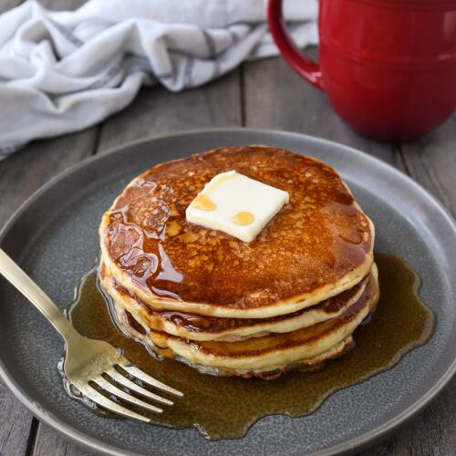 A plate with a stack of packcakes with a pat of butter and drizzeld with syrup. A red coffee cup and a towel in the background.