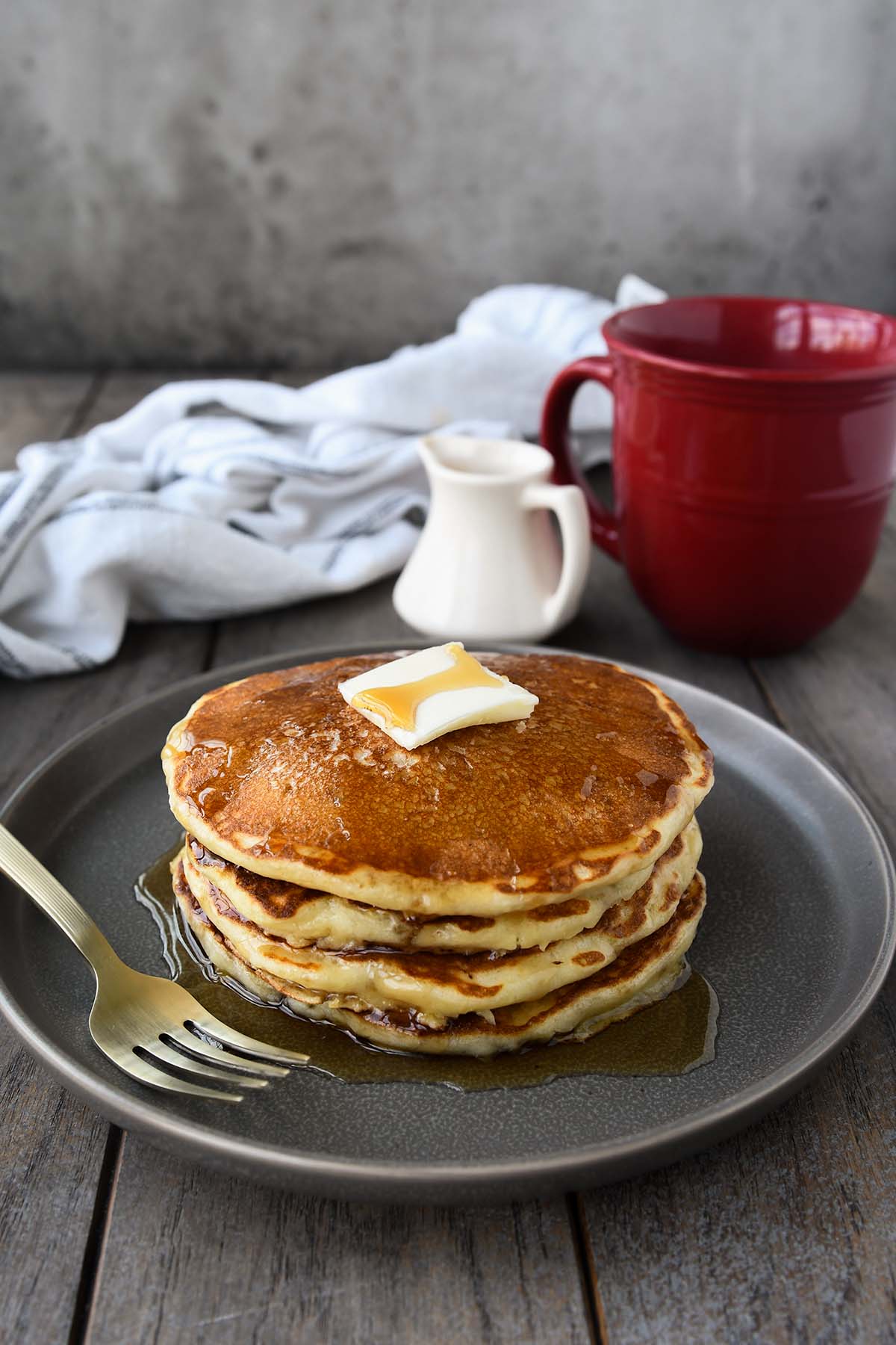 A plate with a stack of packcakes with a pat of butter and drizzeld with syrup. A red coffee cup and a towel in the background.