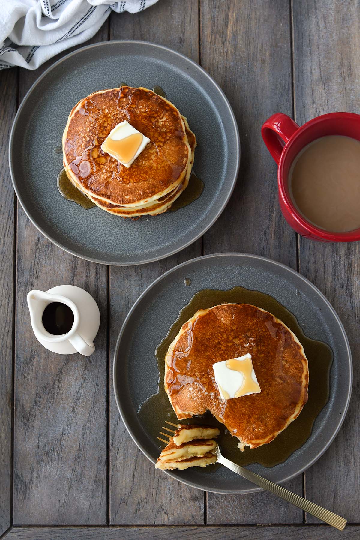 Above view of two plates of pancakes with a red coffee cup, a white towel, and a little container of syrup.