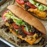 Chopped sandwiches overflowing with beef, cheese, lettuce, and tomato all lined up on a baking sheet with a striped towel in background.