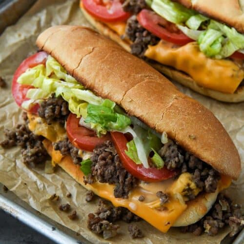 Chopped sandwiches overflowing with beef, cheese, lettuce, and tomato all lined up on a baking sheet with a striped towel in background.