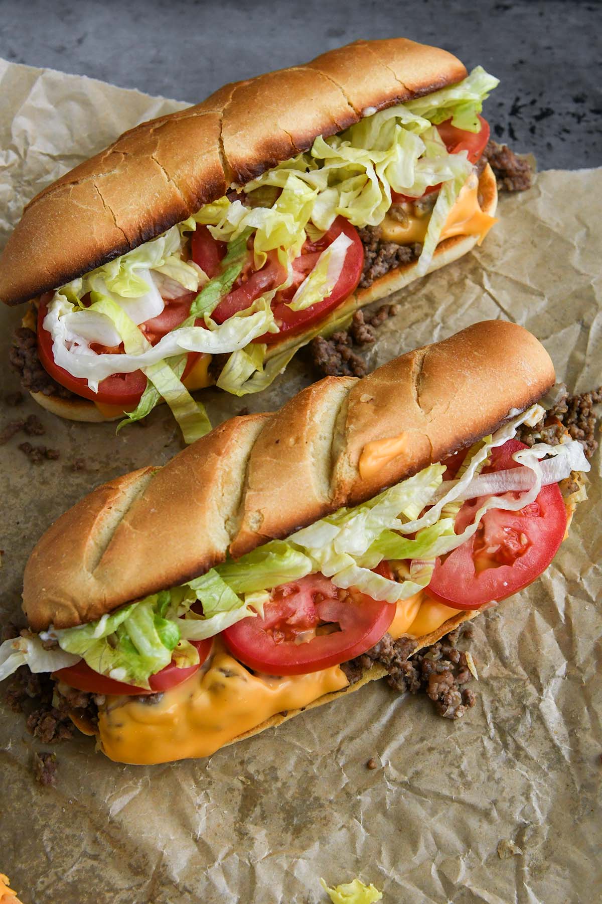 Two chopped cheese sandwiches overflowing with beef, cheese, lettuce, and tomato on a piece of parchment paper.