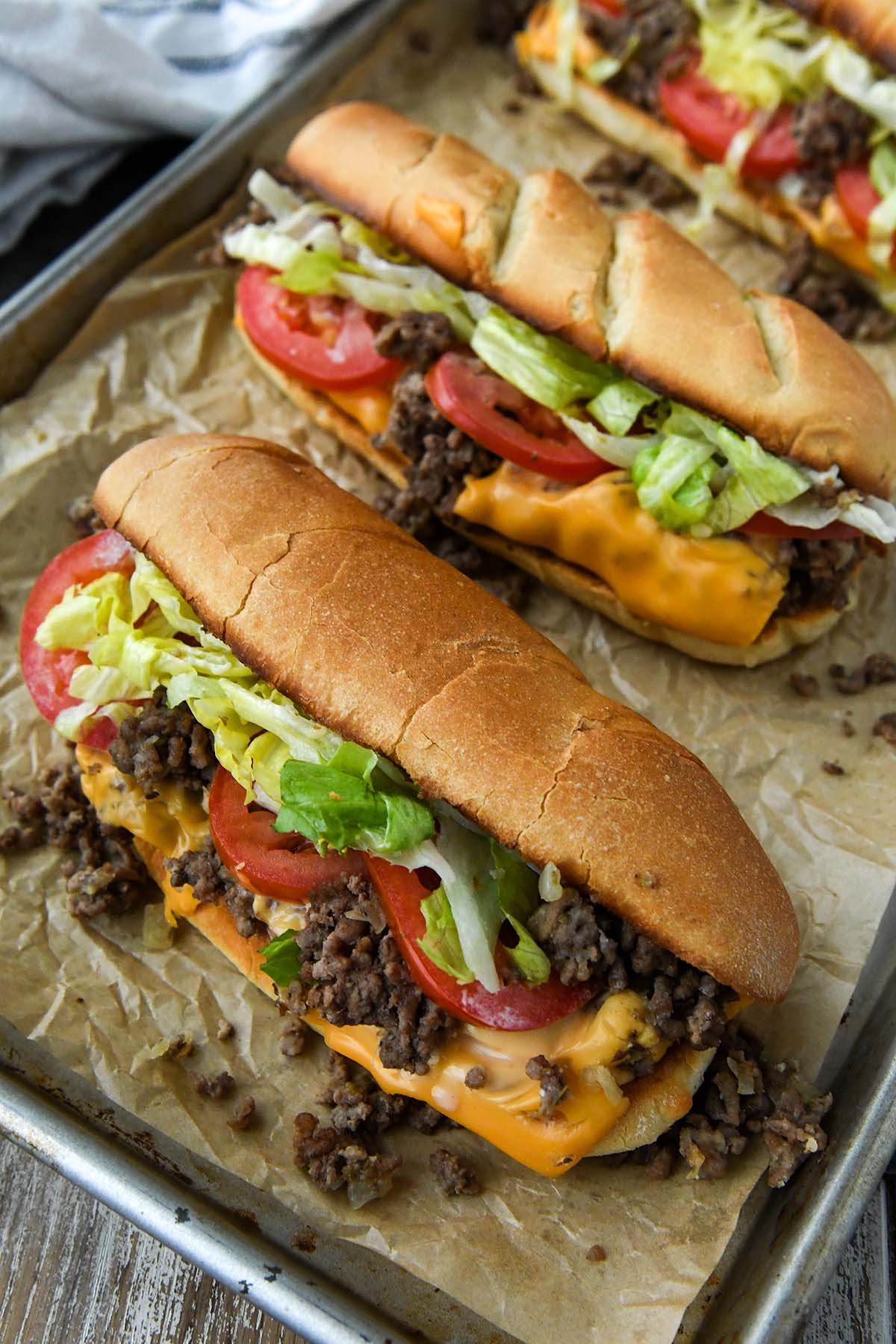 Chopped sandwiches overflowing with beef, cheese, lettuce, and tomato all lined up on a baking sheet with a striped towel in background.