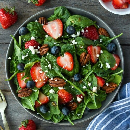 Above view of a plate full of spinach salad topped with strawberries, blueberries, nuts, feta cheese and balsamic dressing. Strawberries, blue striped towel and gold forks are around the plate.