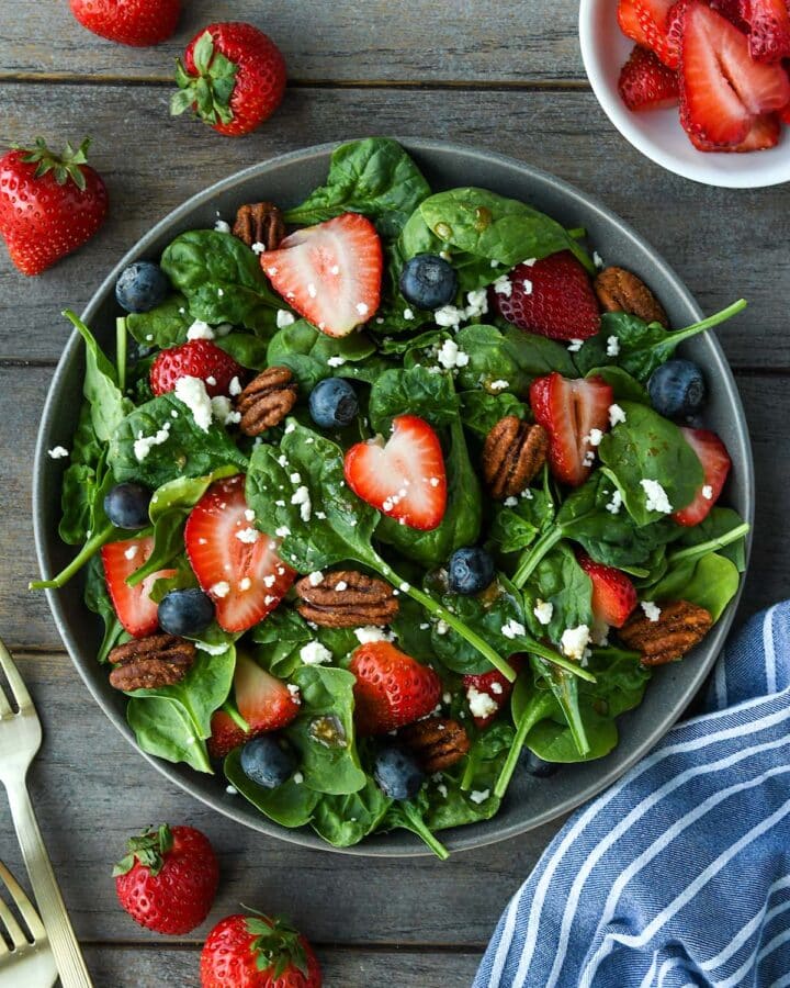 Above view of a plate full of spinach salad topped with strawberries, blueberries, nuts, feta cheese and balsamic dressing. Strawberries, blue striped towel and gold forks are around the plate.