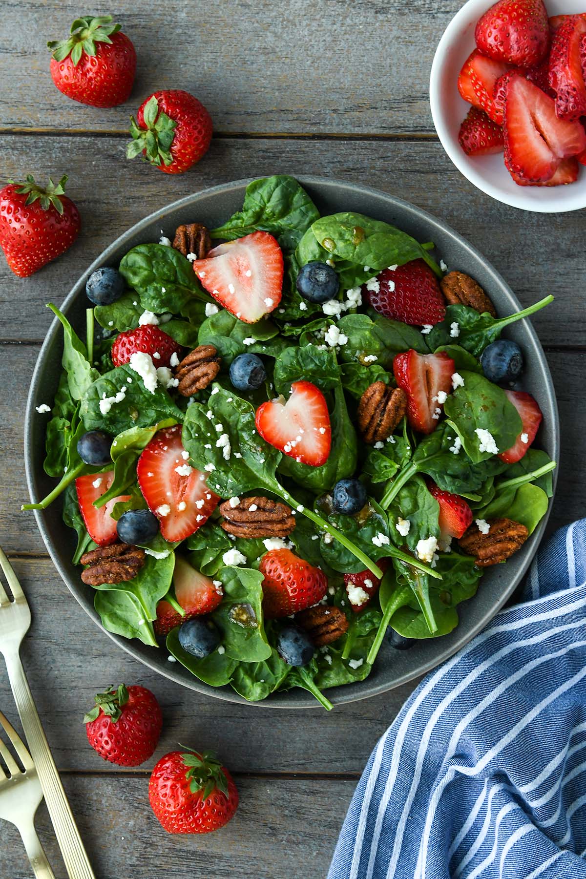 Above view of a plate full of spinach salad topped with strawberries, blueberries, nuts, feta cheese and balsamic dressing. Strawberries, blue striped towel and gold forks are around the plate.