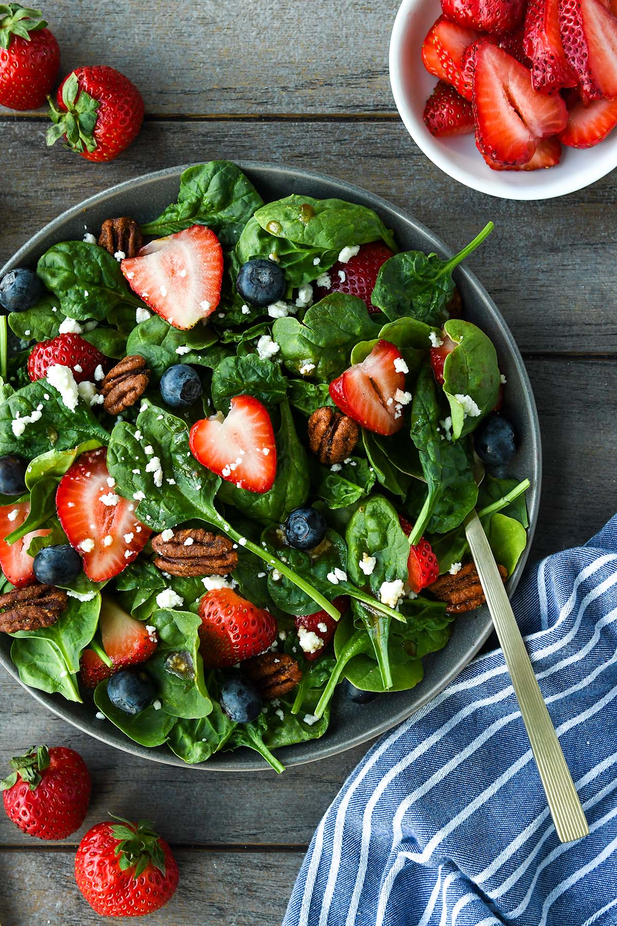Above view of a plate full of spinach salad topped with strawberries, blueberries, nuts, feta cheese and balsamic dressing. Strawberries, blue striped towel and gold forks are around the plate.