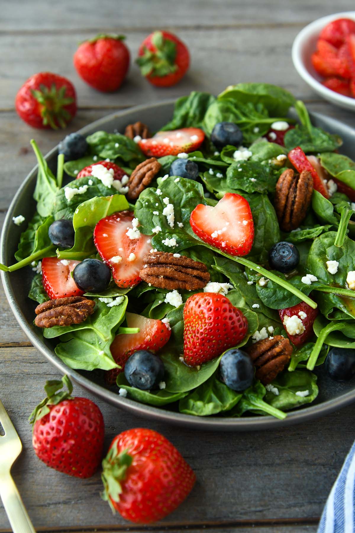 A plate full of spinach salad topped with strawberries, blueberries, nuts, feta cheese and balsamic dressing. Strawberries, blue striped towel and gold forks are around the plate.