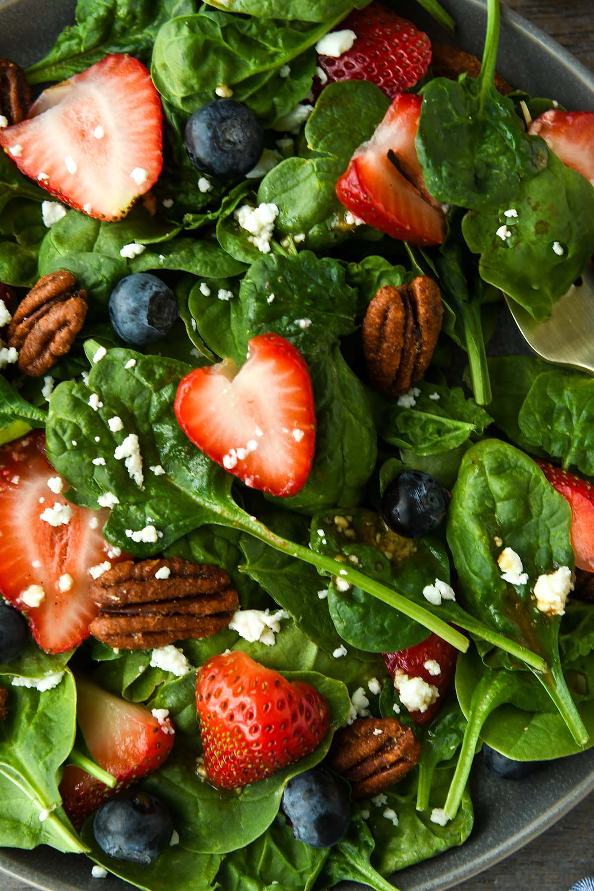 Very up close view of a plate full of spinach salad topped with strawberries, blueberries, nuts, feta cheese and balsamic dressing. Strawberries, blue striped towel and gold forks are around the plate.