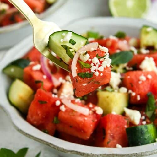 A bite of watermelon salad on a fork above a full bowl with a sprig of mint and half of a lime.
