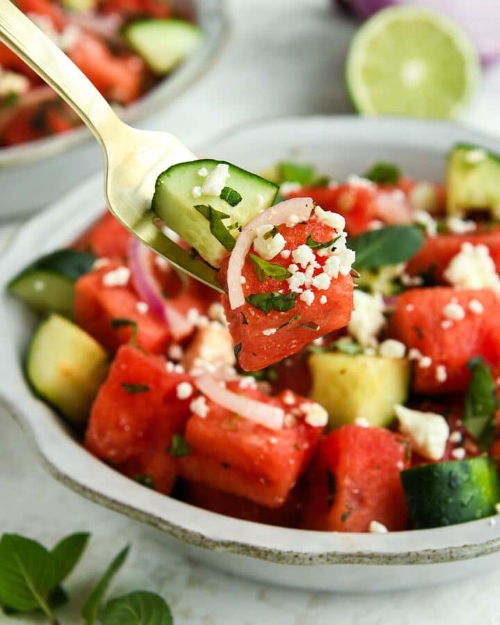 A bite of watermelon salad on a fork above a full bowl with a sprig of mint and half of a lime.