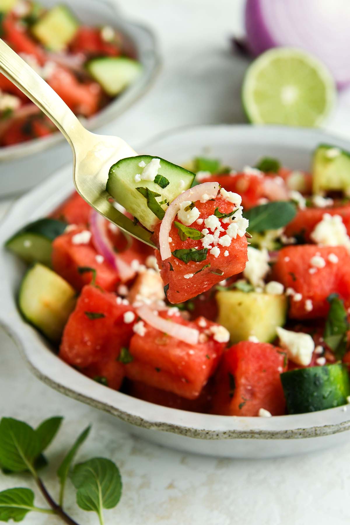 A bite of watermelon salad on a fork above a full bowl with a sprig of mint and half of a lime.