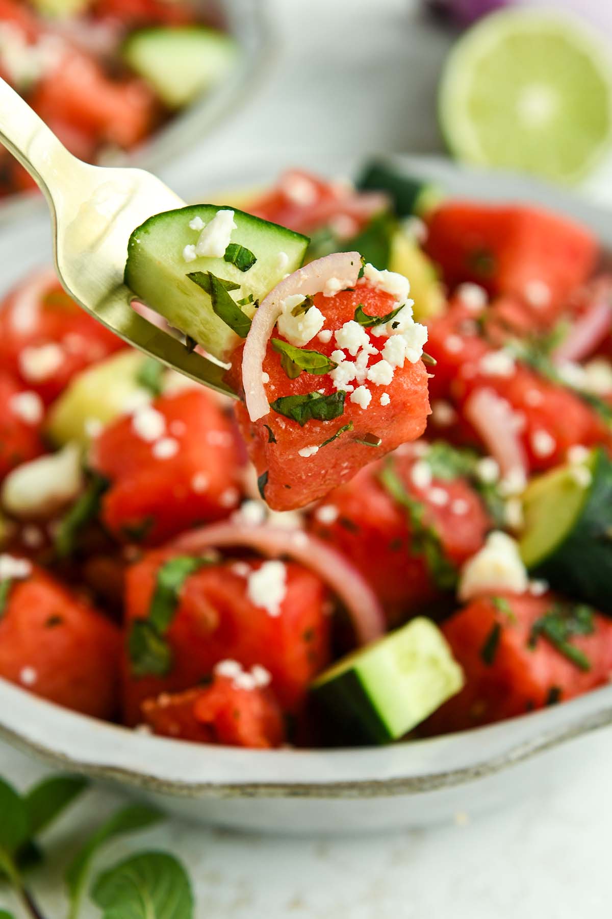 A bite of watermelon salad on a fork above a full bowl with a sprig of mint and half of a lime.