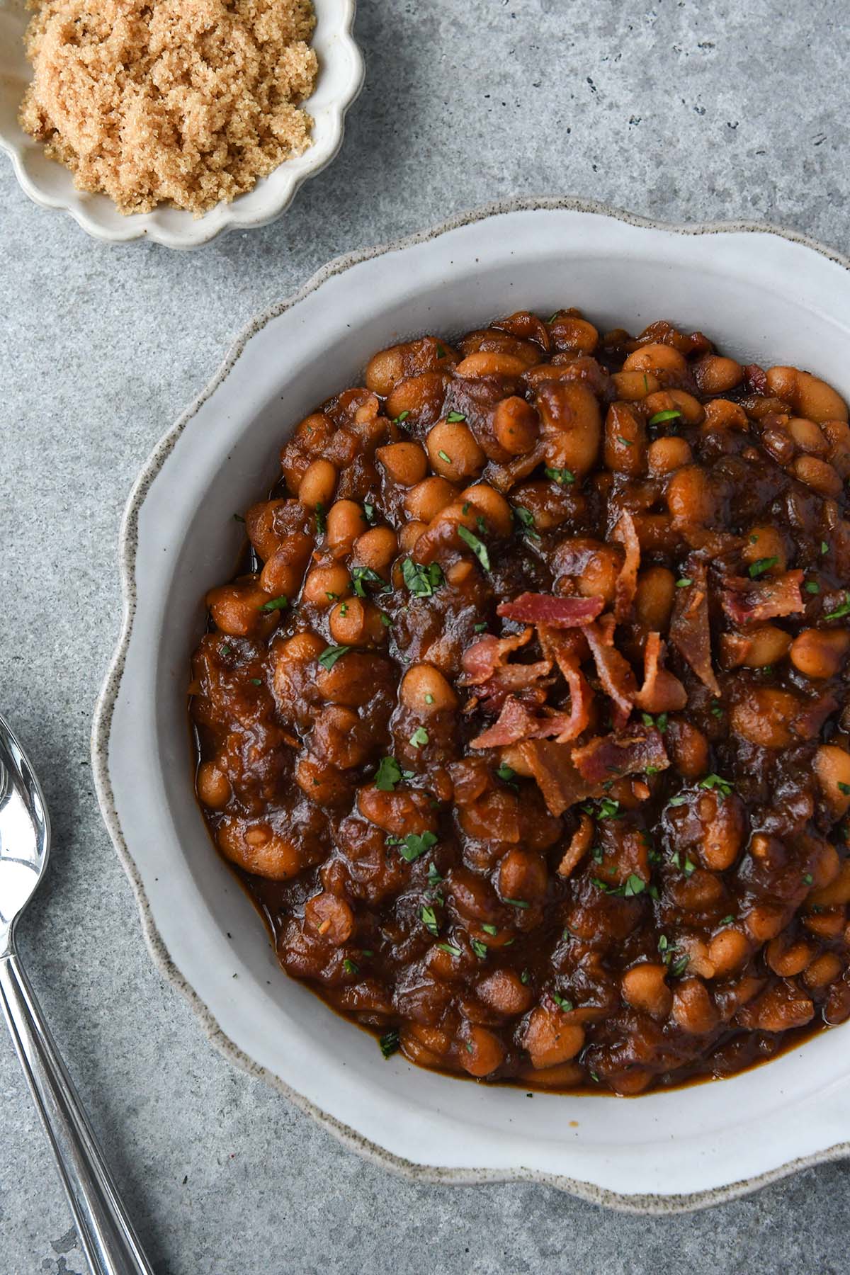 Up close and above view of a serving bowl of bbq baked beans, small dish of brown sugar, and a spoon.