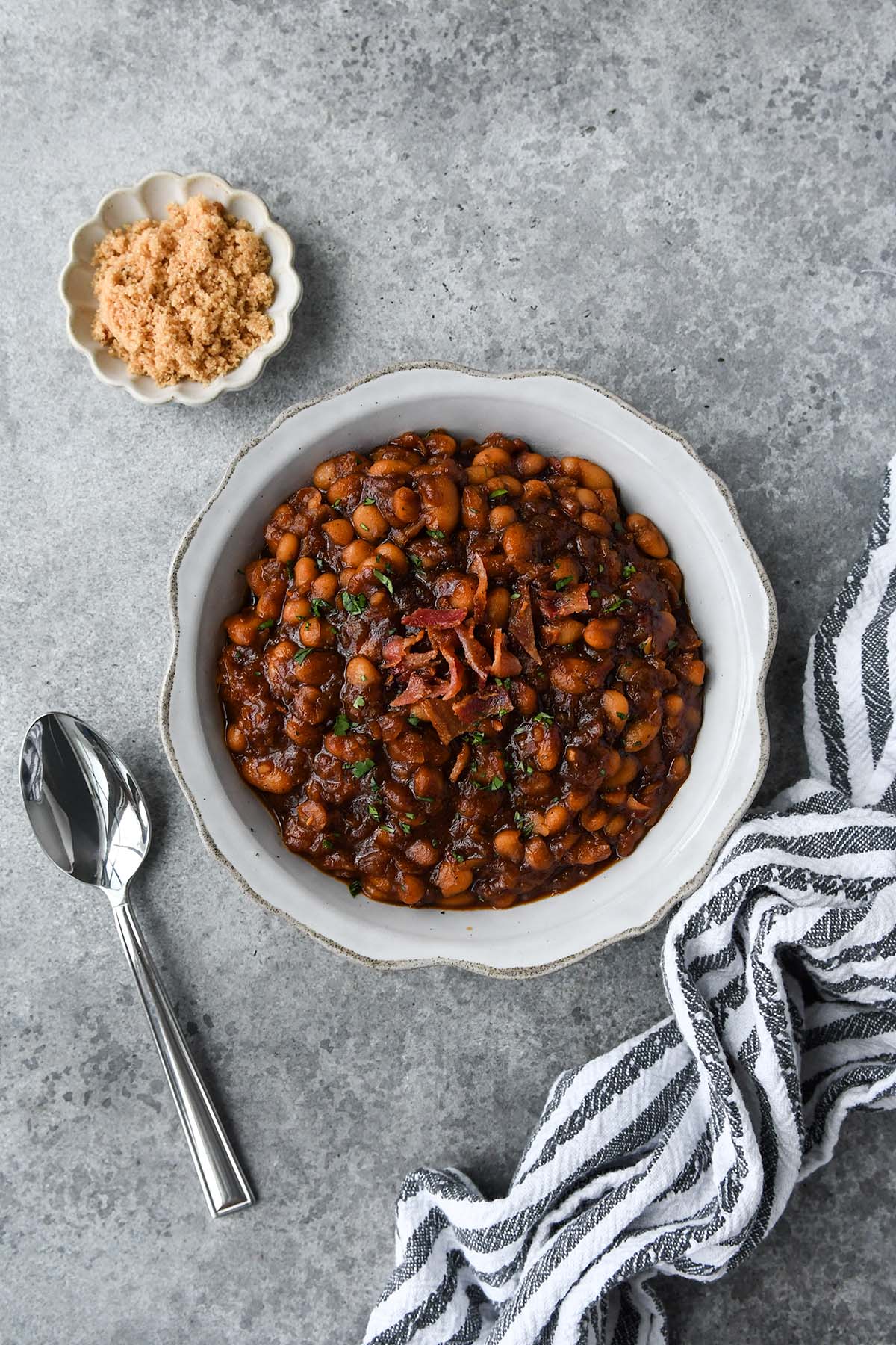 Above view of a serving bowl of bbq baked beans, a striped towel, and a small dish of brown sugar.