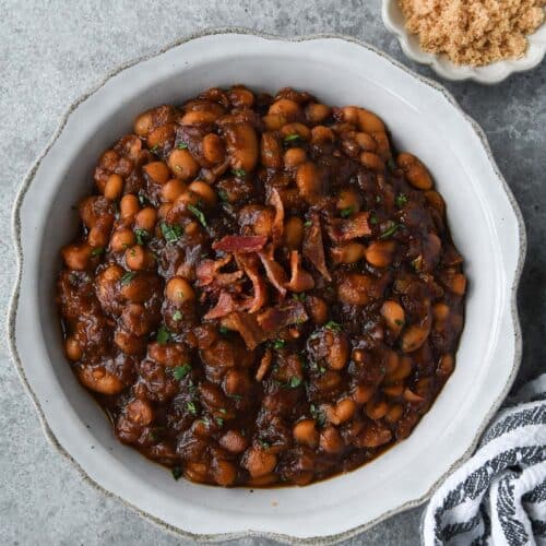 Above view of a serving bowl of bbq baked beans, a striped towel, and a small dish of brown sugar.