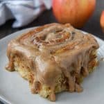 Up close of an apple pie cinnamon roll dripping with spiced frosting on a plate with apples and a towel in the background.