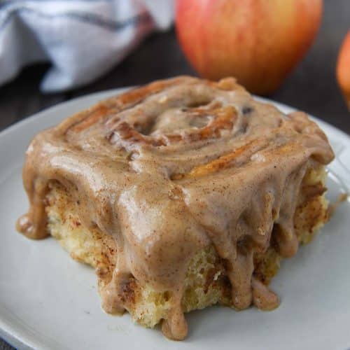 Up close of an apple pie cinnamon roll dripping with spiced frosting on a plate with apples and a towel in the background.