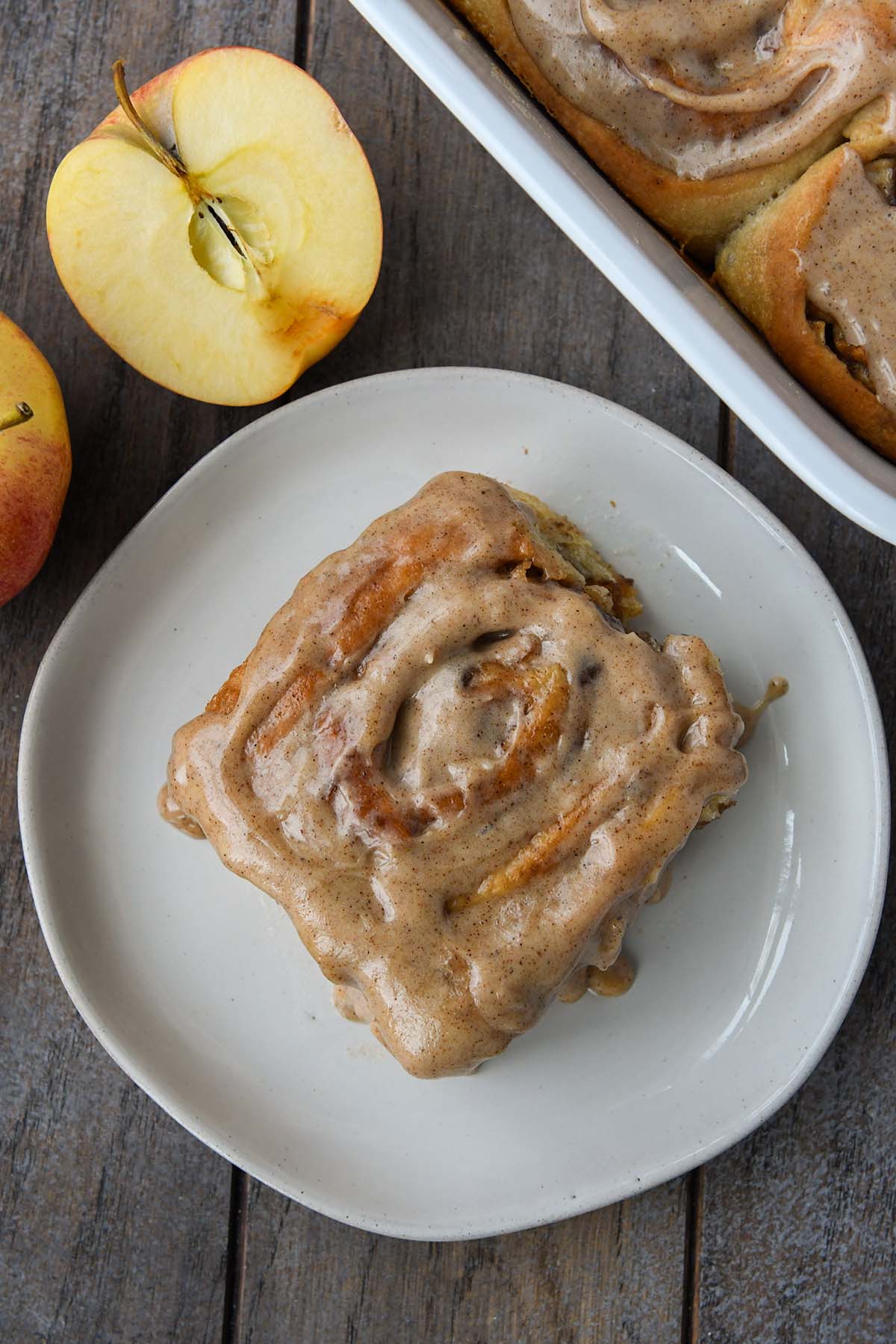 Overhead shot of an apple cinnamon roll on a plate with a sliced apple and tray of more rolls.
