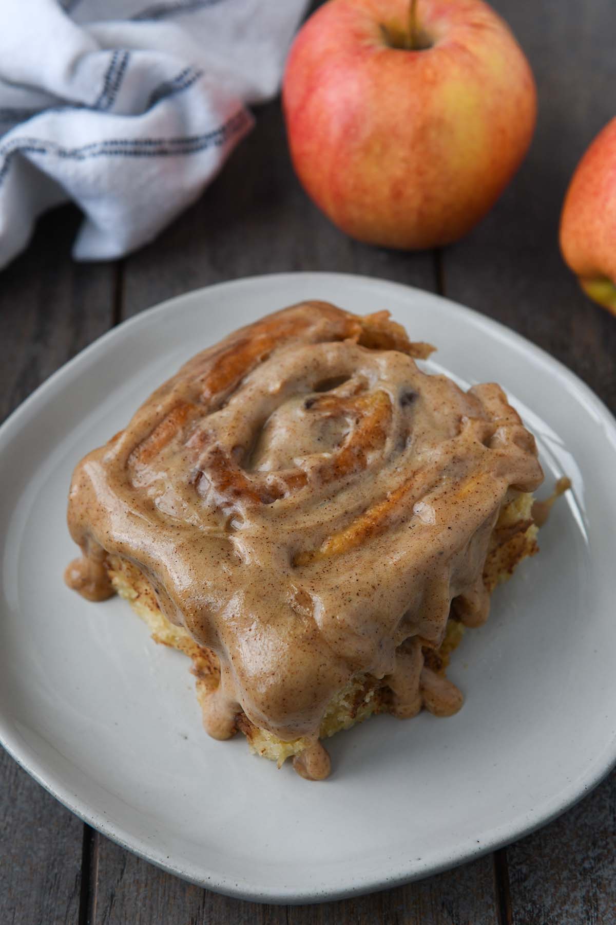 Up close of an apple pie cinnamon roll dripping with spiced frosting on a plate with apples and a towel in the background.
