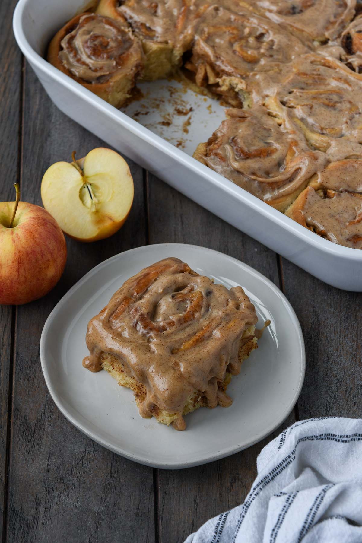 Up close of an apple pie cinnamon roll dripping with spiced frosting on a plate with apples and a tray of more rolls in the background.