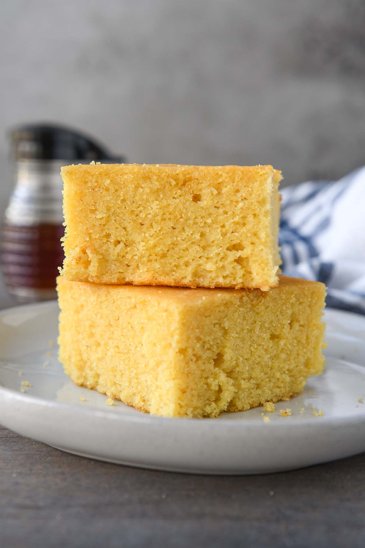 Two stacked pieces of cornbread on a white plate with a towel and a small bottle of honey is in the background.